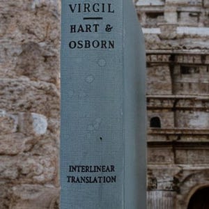 May include: A vintage book with a faded blue cover. The title reads "VIRGIL HART & OSBORN" with "INTERLINEAR TRANSLATION" below. The book's spine is visible against a backdrop of weathered stone and architectural details.