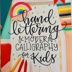May include: A book titled "Hand Lettering & Modern Calligraphy for Kids" with a rainbow sticker and various art supplies. The cover features the title in elegant script. The book includes over 15 project and party ideas.