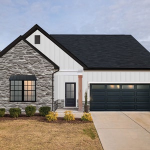 May include: A white and gray farmhouse style home with a black roof, black garage door, and a black front door. The home has a stone facade and a small front yard with bushes and flowers.