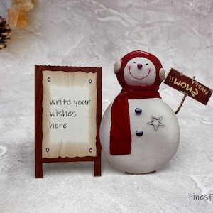 Pu&ograve; includere: Un pupazzo di neve con una sciarpa rossa e un cartello che dice "Buon Natale!!" si trova accanto a un cartello di legno che dice "Scrivi i tuoi desideri qui".