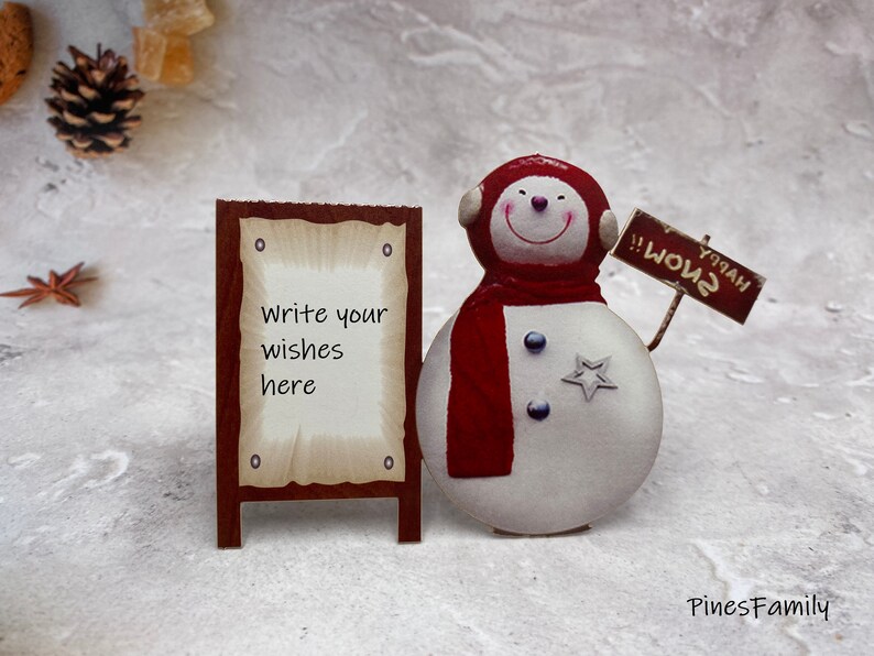 Pu&ograve; includere: Un pupazzo di neve con una sciarpa rossa e un cartello che dice "Buon Natale!!" si trova accanto a un cartello di legno che dice "Scrivi i tuoi desideri qui".