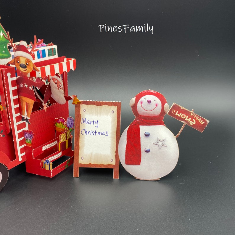 Pu&ograve; includere: Un biglietto di auguri pop-up natalizio rosso e bianco con un pupazzo di neve che tiene un cartello con su scritto "Buone Feste!" Un cartello accanto al pupazzo di neve dice "Buon Natale".
