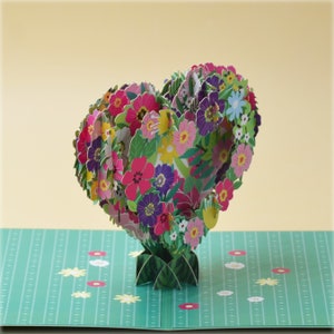 May include: A 3D pop-up card featuring a heart-shaped bouquet of colorful flowers. The flowers are pink, purple, yellow, and white. The card is open and the flowers are standing up.