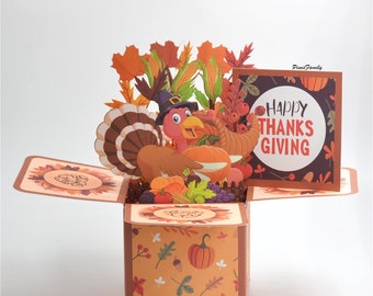 May include: A 3D pop-up card with a Thanksgiving theme. The card features a turkey with a pilgrim hat, a cornucopia, and a variety of fall fruits and vegetables. The card is open and the pop-up is extended. The card has a brown background with a pattern of fall leaves and pumpkins. The card also has a small card attached to the front that says "Happy Thanksgiving".