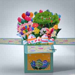May include: A pop-up Easter card featuring a festive scene with colorful Easter eggs, a bunny, flowers, and balloons. The card has the words "Happy Easter" on a sign. The card is in a box with a yellow and green design.
