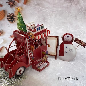 Pu&ograve; includere: Un biglietto pop-up rosso con un camioncino a tema natalizio con una renna, un albero di Natale e dei regali. Accanto al camioncino c'&egrave; un pupazzo di neve con una sciarpa rossa e un cartello che dice "Buon Natale!"