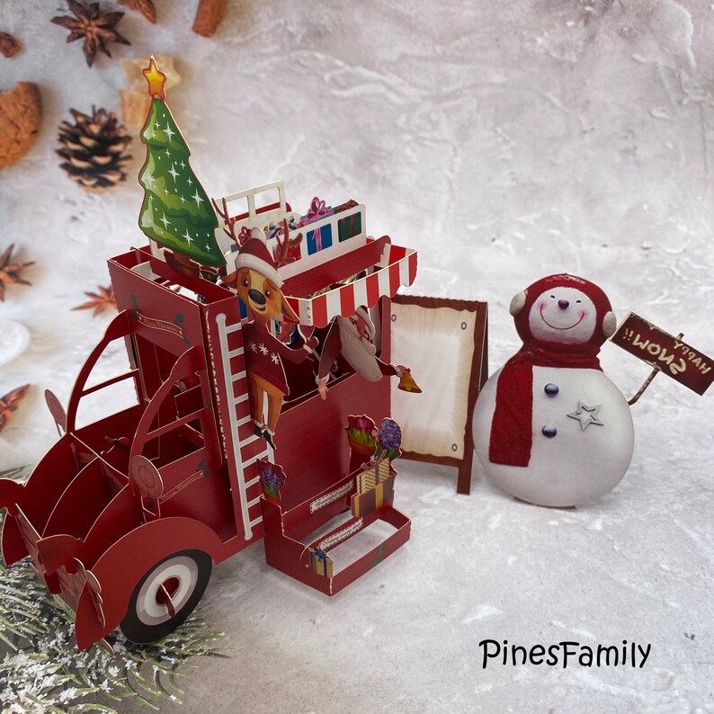 Pu&ograve; includere: Un biglietto pop-up rosso con un camioncino a tema natalizio con una renna, un albero di Natale e dei regali. Accanto al camioncino c'&egrave; un pupazzo di neve con una sciarpa rossa e un cartello che dice "Buon Natale!"