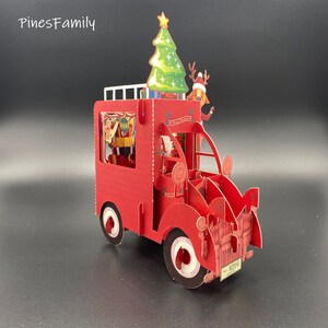 Pu&ograve; includere: Un biglietto pop-up 3D rosso che rappresenta un camion di consegna a tema natalizio con un albero di Natale in cima. Il camion ha un portapacchi bianco sul tetto, un volante bianco e una targa bianca che recita "No. 123456".