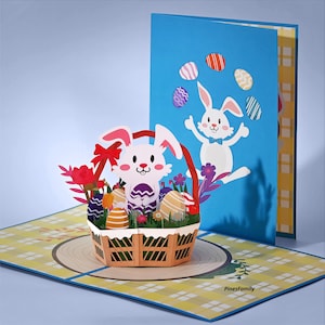 May include: A 3D Easter pop-up card featuring a white bunny in a basket with colorful eggs and flowers. The card has a blue background with a bunny and eggs. The card is open, revealing a yellow and blue patterned surface.