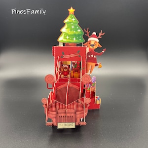 Pu&ograve; includere: Un camioncino rosso vintage con un albero di Natale in cima, una renna sul lato e un orsacchiotto nella cabina. Il camioncino ha una targa che recita "N.J. W2UHG GARDEN STATE".