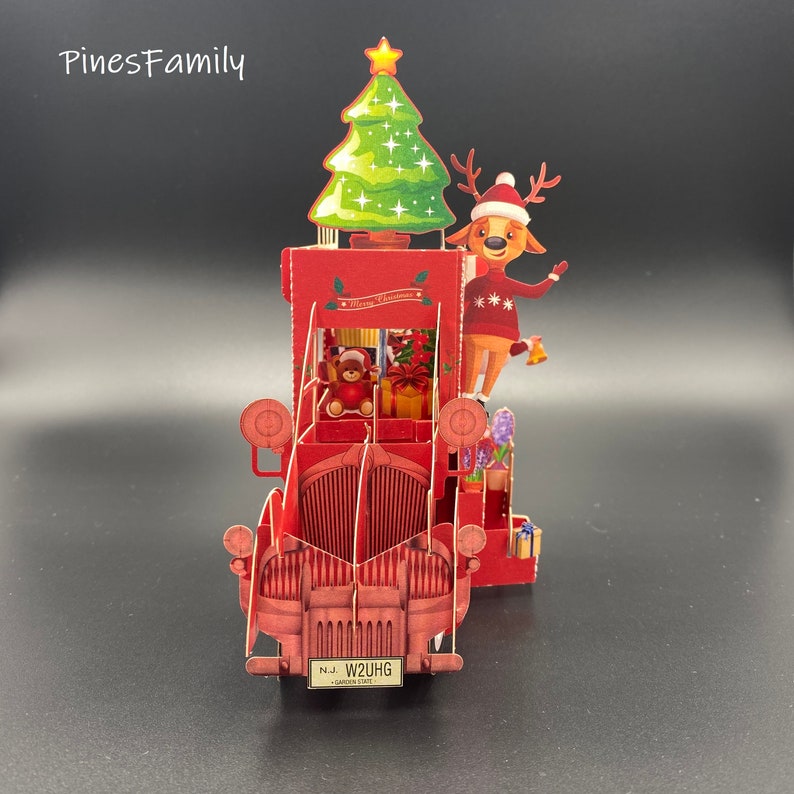 Pu&ograve; includere: Un camioncino rosso vintage con un albero di Natale in cima, una renna sul lato e un orsacchiotto nella cabina. Il camioncino ha una targa che recita "N.J. W2UHG GARDEN STATE".