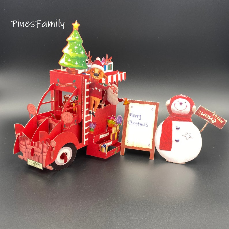 Pu&ograve; includere: Un camioncino rosso con un albero di Natale in cima, una renna che sale su una scala e un pupazzo di neve che tiene un cartello con su scritto "Happy Snow". Il camioncino ha un cartello con su scritto "Merry Christmas".