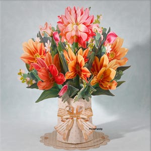 May include: A 3D pop-up floral arrangement featuring orange, red, and pink flowers. The bouquet is in a beige vase with a bow and polka dots. The base is a scalloped edge circle. The text "PinesFamily" is visible.