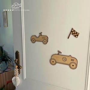 Peut inclure: Trois découpes murales en bois clair représentant deux voitures de course de style vintage et un drapeau à damier. Une voiture est plus petite, tandis que la plus grande affiche le numéro 18 sur son flanc. Ces formes en bois sont fixées sur une porte.
