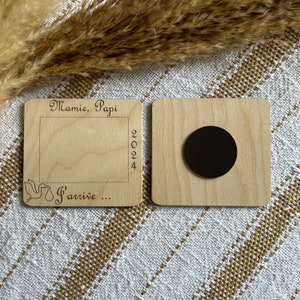 May include: Two wooden magnets. One has a frame with the words "Mamie, Papi" and "2024" engraved. The other magnet has a black circle. The text "S'arrive..." is also engraved on the first magnet.