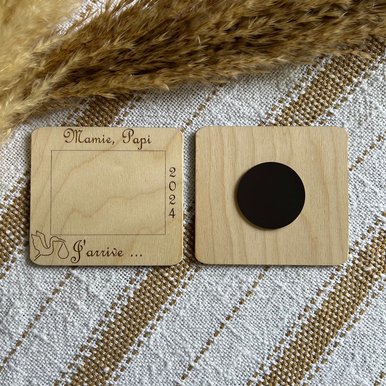 May include: Two wooden magnets. One has a frame with the words "Mamie, Papi" and "2024" engraved. The other magnet has a black circle. The text "S'arrive..." is also engraved on the first magnet.