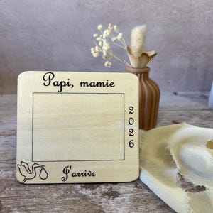 May include: A wooden announcement plaque with the words "Papi, mamie" and "J'arrive" in script. The year "2026" is printed vertically. A stork illustration is in the lower left corner. A small vase with dried flowers is in the background.