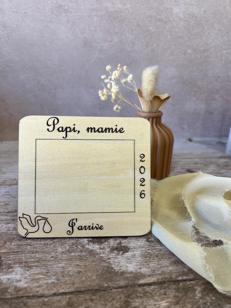 May include: A wooden announcement plaque with the words "Papi, mamie" and "J'arrive" in script. The year "2026" is printed vertically. A stork illustration is in the lower left corner. A small vase with dried flowers is in the background.