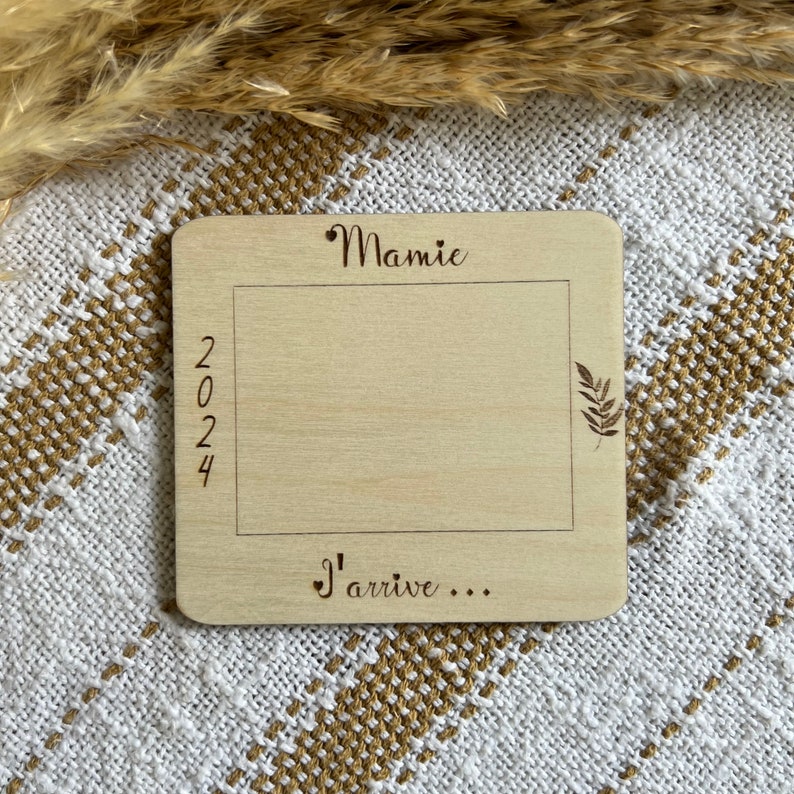 May include: A light-colored wooden plaque with the word "Mamie" at the top and "2024" on the left side. The phrase "J'arrive..." is at the bottom. A rectangular frame is etched into the wood, with a leaf design on the right.