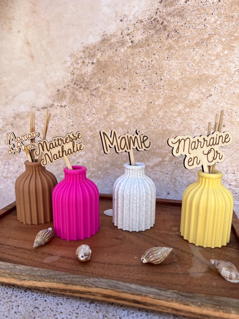 May include: Four small, ribbed, cylindrical vases in brown, hot pink, white speckled, and yellow. Each vase holds wooden sticks with laser-cut wooden toppers featuring the French text Maman d'Amour, Ma&icirc;tresse Nathalie, Mamie, and Marraine en Or.