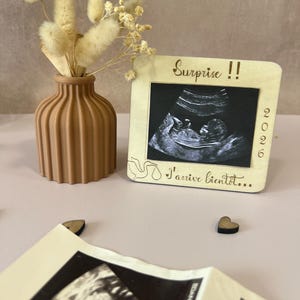 May include: A wooden frame with a sonogram image, the text "Surprise !!" and "I'arrive bient&ocirc;t..." is displayed. A brown vase with dried flowers is next to the frame. Two small wooden hearts are also visible.