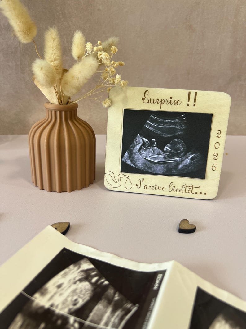 May include: A wooden frame with a sonogram image, the text "Surprise !!" and "I'arrive bient&ocirc;t..." is displayed. A brown vase with dried flowers is next to the frame. Two small wooden hearts are also visible.
