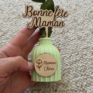 May include: A light green ribbed vase featuring a wooden topper with the text Bonne f&ecirc;te Maman and a circular wooden tag engraved with a flower icon and the text Maman Ch&eacute;rie, containing a single green eucalyptus stem.