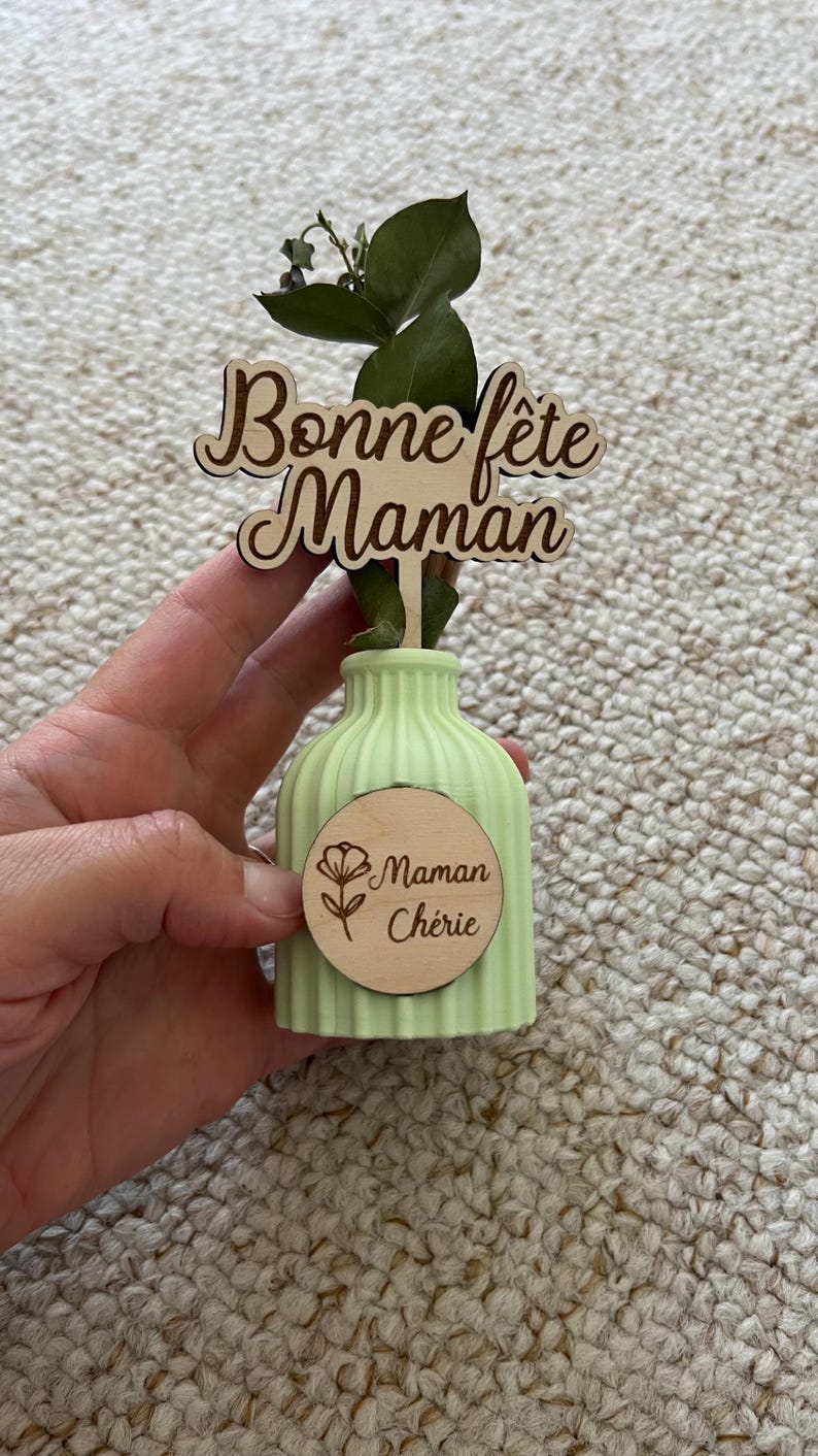 May include: A light green ribbed vase featuring a wooden topper with the text Bonne f&ecirc;te Maman and a circular wooden tag engraved with a flower icon and the text Maman Ch&eacute;rie, containing a single green eucalyptus stem.