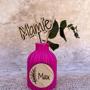 May include: A bright pink ribbed vase featuring a circular wooden plaque with the name Max and a leaf illustration. A wooden cutout with the word Mamie is inserted into the vase alongside a small green leafy branch, creating a decorative display.