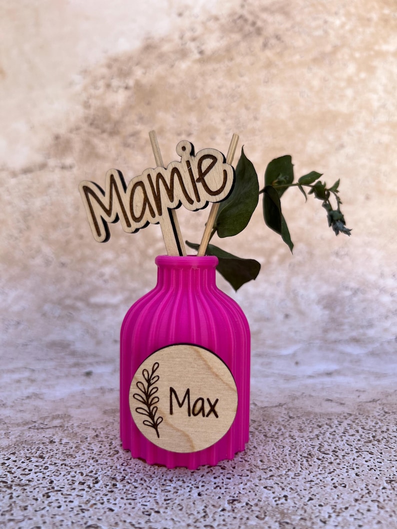 May include: A bright pink ribbed vase featuring a circular wooden plaque with the name Max and a leaf illustration. A wooden cutout with the word Mamie is inserted into the vase alongside a small green leafy branch, creating a decorative display.