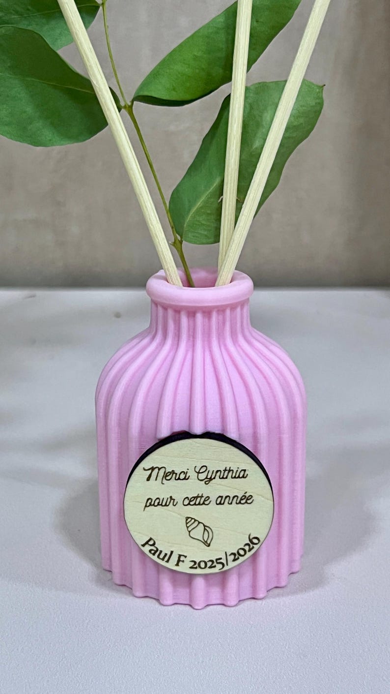 May include: A light pink ribbed diffuser bottle with three wooden reeds and green leaves. A circular wooden tag is attached to the front, featuring the text "Merci Cynthia pour cette ann&eacute;e", a small seashell icon, and the text "Paul F 2025/2026".