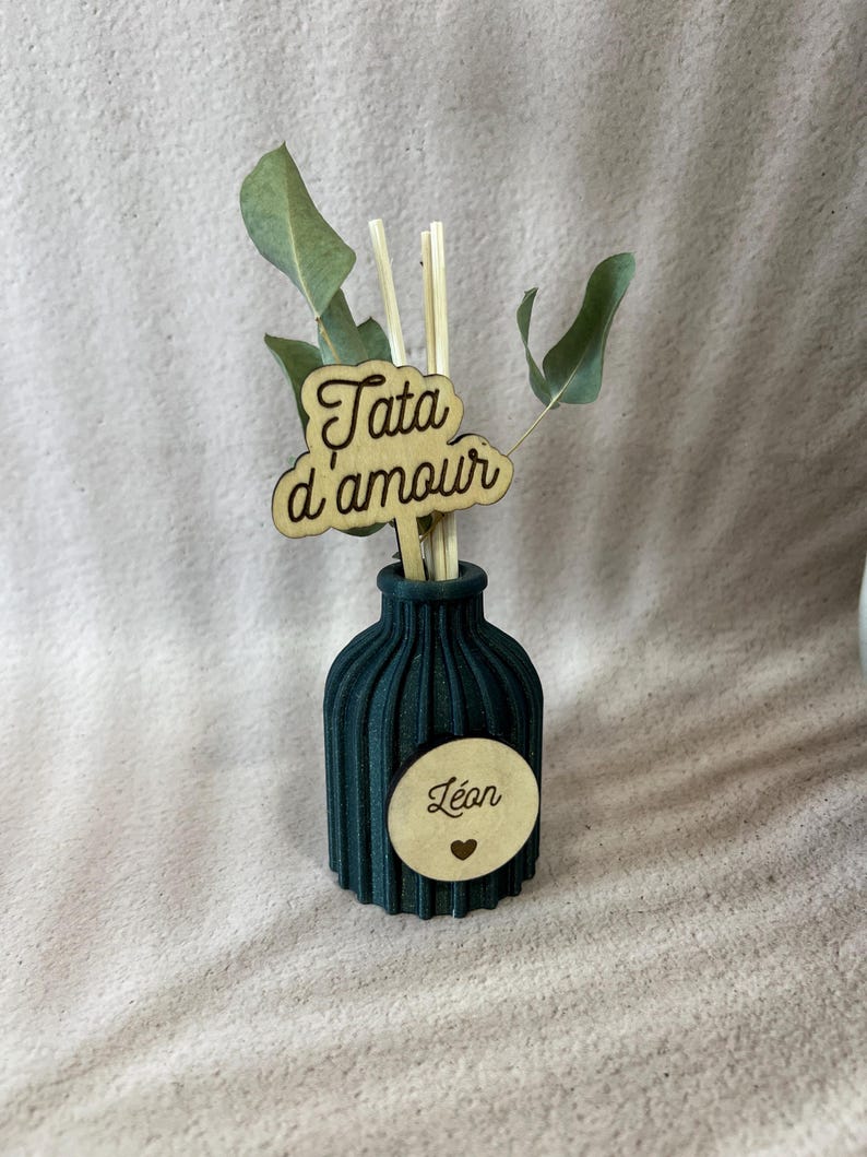 May include: A dark green ribbed diffuser bottle featuring a circular wooden tag with the name L&eacute;an and a small heart icon. A wooden topper with the text Tata d'amour is inserted into the bottle along with several reed sticks and green leaves.