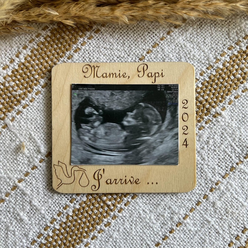 May include: A wooden frame with an ultrasound image, the text "Mamie, Papi" and "I'arrive..." is engraved on the frame. The year "2024" is also engraved on the frame. A stork illustration is also engraved on the frame.