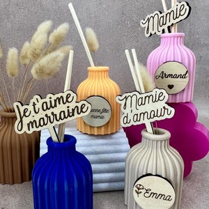 May include: A collection of ribbed, colorful vases in blue, orange, pink, beige, and brown, each containing wooden sticks and decorative dried grass. Wooden plaques are attached, featuring text such as Je t'aime marraine, Mamie, Mamie d'amour, Armand, Emma, and Bonne f&ecirc;te mamie.