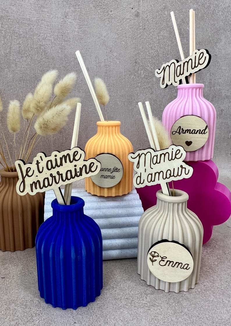 May include: A collection of ribbed, colorful vases in blue, orange, pink, beige, and brown, each containing wooden sticks and decorative dried grass. Wooden plaques are attached, featuring text such as Je t'aime marraine, Mamie, Mamie d'amour, Armand, Emma, and Bonne f&ecirc;te mamie.