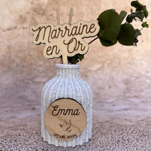 May include: A white speckled ribbed ceramic vase containing a green eucalyptus sprig. A wooden cutout topper reads "Marraine en Or" and a circular wooden plaque on the front features the name "Emma," a dove illustration, and the date "07.06.2025."