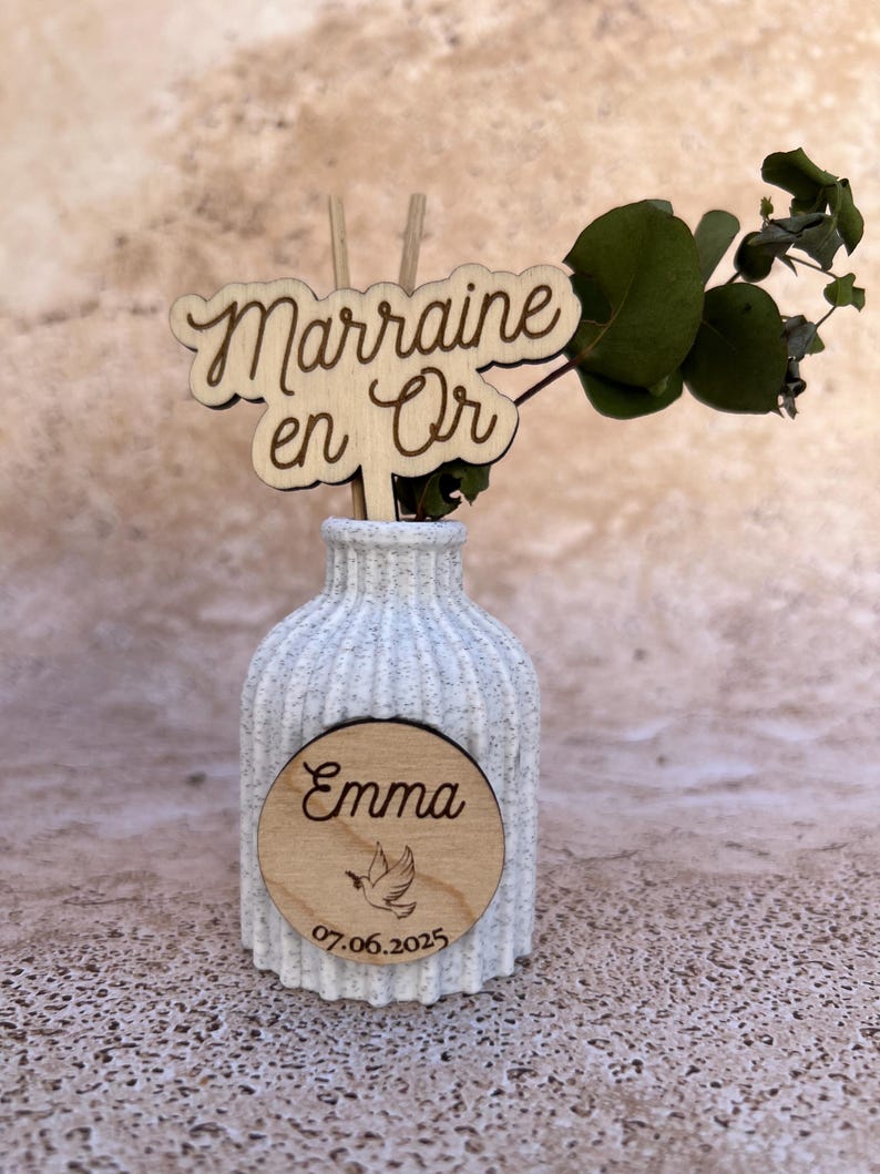May include: A white speckled ribbed ceramic vase containing a green eucalyptus sprig. A wooden cutout topper reads "Marraine en Or" and a circular wooden plaque on the front features the name "Emma," a dove illustration, and the date "07.06.2025."