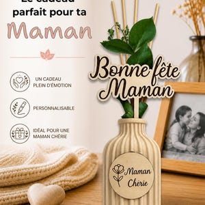 May include: A beige ribbed ceramic diffuser bottle with a wooden plaque reading Maman Ch&eacute;rie and a flower icon. A wooden topper reads Bonne f&ecirc;te Maman, accompanied by green leaves and wooden reeds. Text includes Le cadeau parfait pour ta Maman.