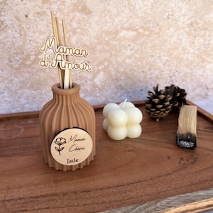 May include: A tan ribbed ceramic diffuser bottle with wooden reeds featuring a cutout sign that reads Maman d'Amour and a circular wooden tag labeled Maman Ch&eacute;rie Jade. Beside it sits a white bubble candle, a pinecone, and a piece of palo santo.