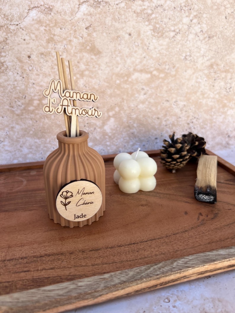 May include: A tan ribbed ceramic diffuser bottle with wooden reeds featuring a cutout sign that reads Maman d'Amour and a circular wooden tag labeled Maman Ch&eacute;rie Jade. Beside it sits a white bubble candle, a pinecone, and a piece of palo santo.
