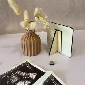 May include: A neutral-toned, ribbed vase holds dried flowers, next to a wooden photo frame and a small wooden heart. Ultrasound images are in the foreground. The items are arranged on a light pink surface, creating a soft, natural aesthetic.