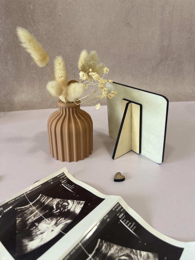 May include: A neutral-toned, ribbed vase holds dried flowers, next to a wooden photo frame and a small wooden heart. Ultrasound images are in the foreground. The items are arranged on a light pink surface, creating a soft, natural aesthetic.