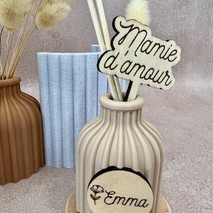 May include: A beige ribbed vase on a wooden base containing two wooden sticks and a wooden sign with the text Mamie d'amour. A circular wooden plaque attached to the front of the vase features a flower icon and the name Emma in cursive script.