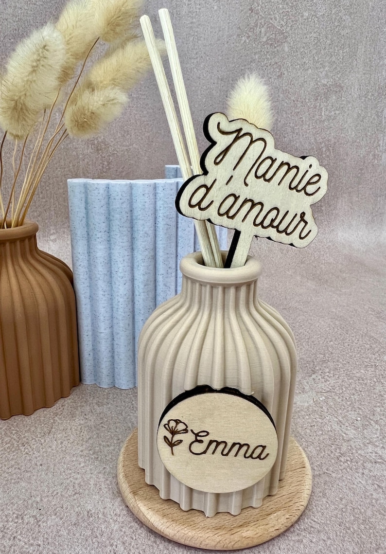 May include: A beige ribbed vase on a wooden base containing two wooden sticks and a wooden sign with the text Mamie d'amour. A circular wooden plaque attached to the front of the vase features a flower icon and the name Emma in cursive script.