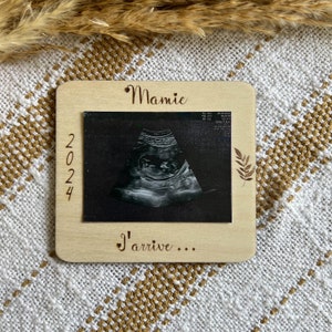 May include: A wooden frame with an ultrasound image. The frame has the word "Mamie" at the top, the year "2024" on the left, and the phrase "I'arrive..." at the bottom. A leaf design is on the right side.