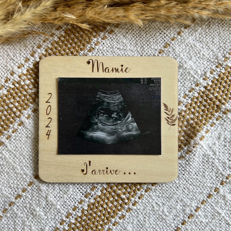 May include: A wooden frame with an ultrasound image. The frame has the word "Mamie" at the top, the year "2024" on the left, and the phrase "I'arrive..." at the bottom. A leaf design is on the right side.