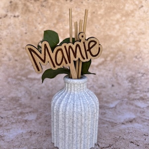 May include: A white speckled ribbed vase containing several thin wooden reeds and a wooden cutout sign with the word Mamie in a cursive script font. A small green leaf sprig is tucked behind the wooden sign inside the vase.