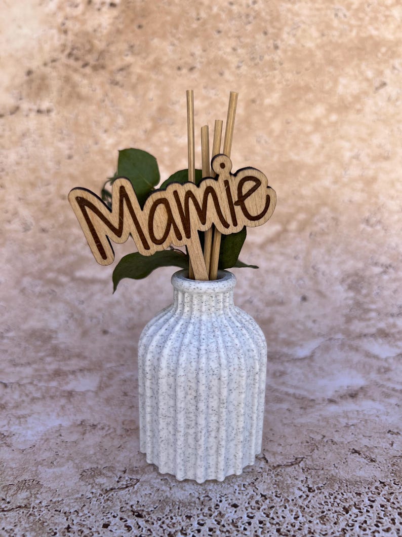 May include: A white speckled ribbed vase containing several thin wooden reeds and a wooden cutout sign with the word Mamie in a cursive script font. A small green leaf sprig is tucked behind the wooden sign inside the vase.