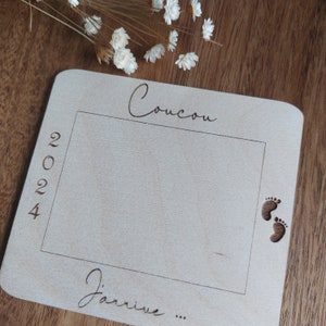 May include: A light-colored wooden baby announcement plaque with the word "Coucou" at the top and "2024" on the left side. The phrase "J'arrive..." is at the bottom, with engraved footprints on the right.