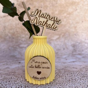 May include: A yellow ribbed vase featuring a circular wooden plaque with the text "Merci pour cette belle ann&eacute;e", a heart icon, and "Jules 2024". A wooden topper displays the text "Ma&icirc;tresse Nathalie" alongside a green eucalyptus sprig.